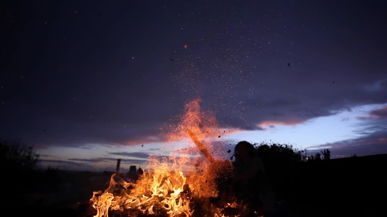Old wooden items and paper are burning in large open fire in a backyard. Person with curly hair adjusts fire with a stick. Sparks and ash fly everywhere. Sun is setting and sky looks very beautiful