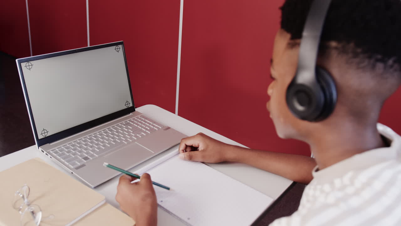 In school, African American teenage boy wearing headphones studying with laptop and notebook at desk