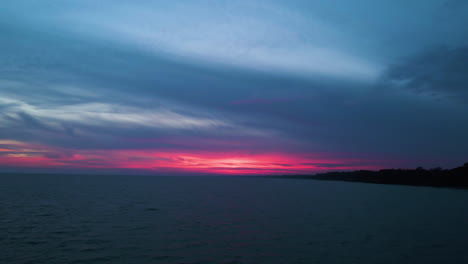 Aerial-trucking-pan-away-from-coastal-land-over-water-as-pink-red-glow-illuminates-horizon