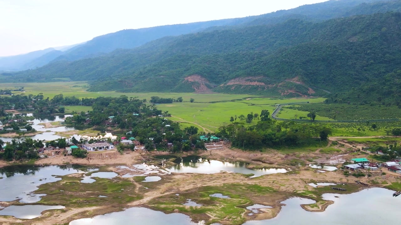 Una tranquilla ripresa aerea di un villaggio rurale di confine accanto a un fiume serpeggiante con campi verdi e montagne sullo sfondo sotto un cielo parzialmente nuvoloso