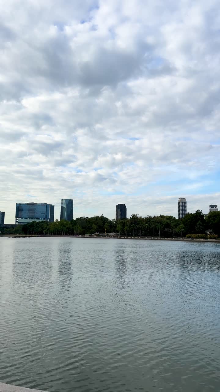 Panorâmica lenta sobre o horizonte da cidade e um parque à beira do lago sob luz diurna nublada, com reflexos calmos na água.