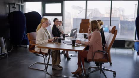 A-group-of-businesswoman-with-an-office-desk-communicate-during-a-meeting-while-their-colleague-a-brunette-girl-in-business-clothes-holds-her-little-infant-daughter-in-her-arms-in-an-office-with-large-windows