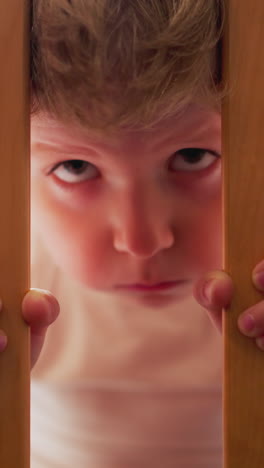 offended little boy looks through wooden grid of baby cradle in children room closeup. upset toddler kid hides in bed. child suffers from loneliness