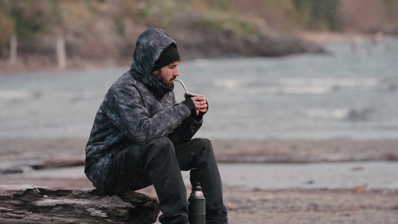 Un uomo con una giacca con cappuccio è seduto su un tronco vicino a un fiume, sorseggiando mate. La scena cattura un momento tranquillo e introspettivo. Adatto per temi di solitudine e riflessione