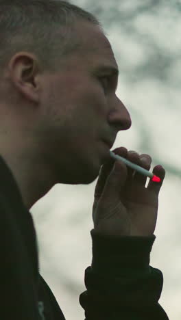 un hombre con una chaqueta verde fumando un cigarrillo al aire libre al anochecer, el fondo está borroso con una luz suave y cálida
