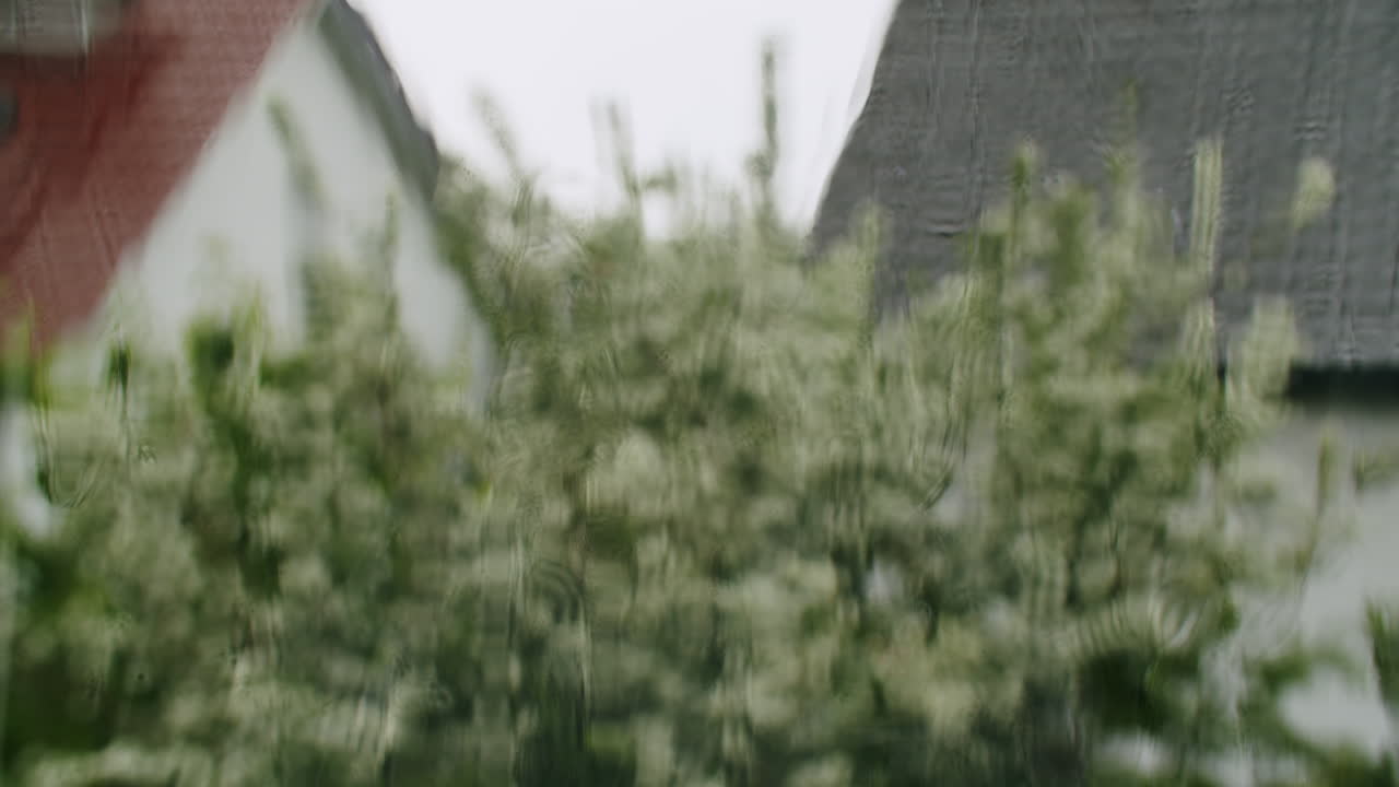 Close-up of slow rain dripping from a balcony edge, captured in soft light. In the background, modest homes, windows, and hedges of a small-town German neighborhood