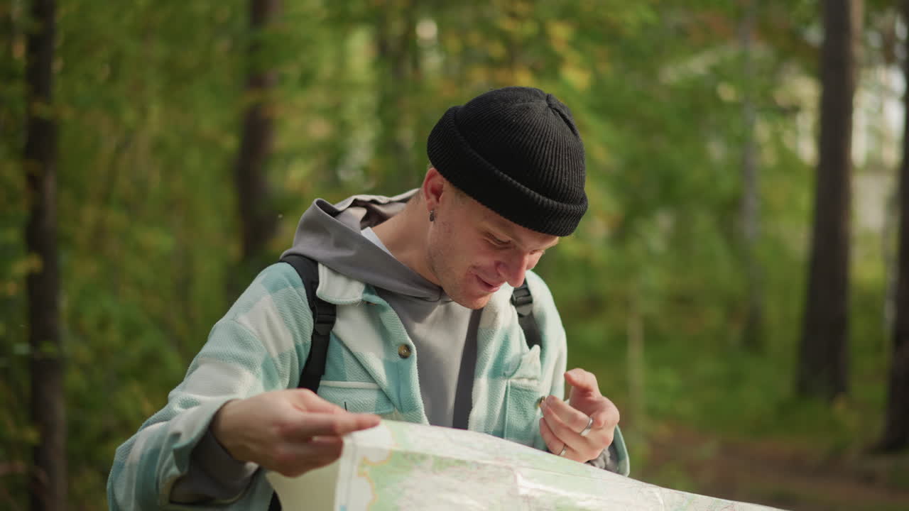 caucasian hiker studying map in forest, puzzled expression, paper map spread wide across hands, backpack straps and knit beanie visible, dappled sunlight through birch canopy, route search