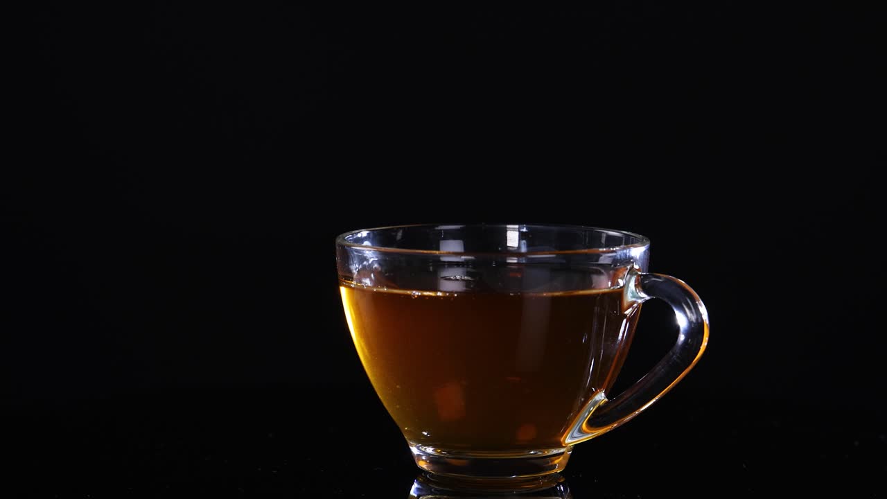 A glass cup filled with tea sits on a black background, showcasing subtle lighting changes and reflections