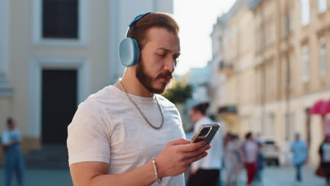 happy joyful young man in wireless headphones choosing listening music dancing outdoors city street