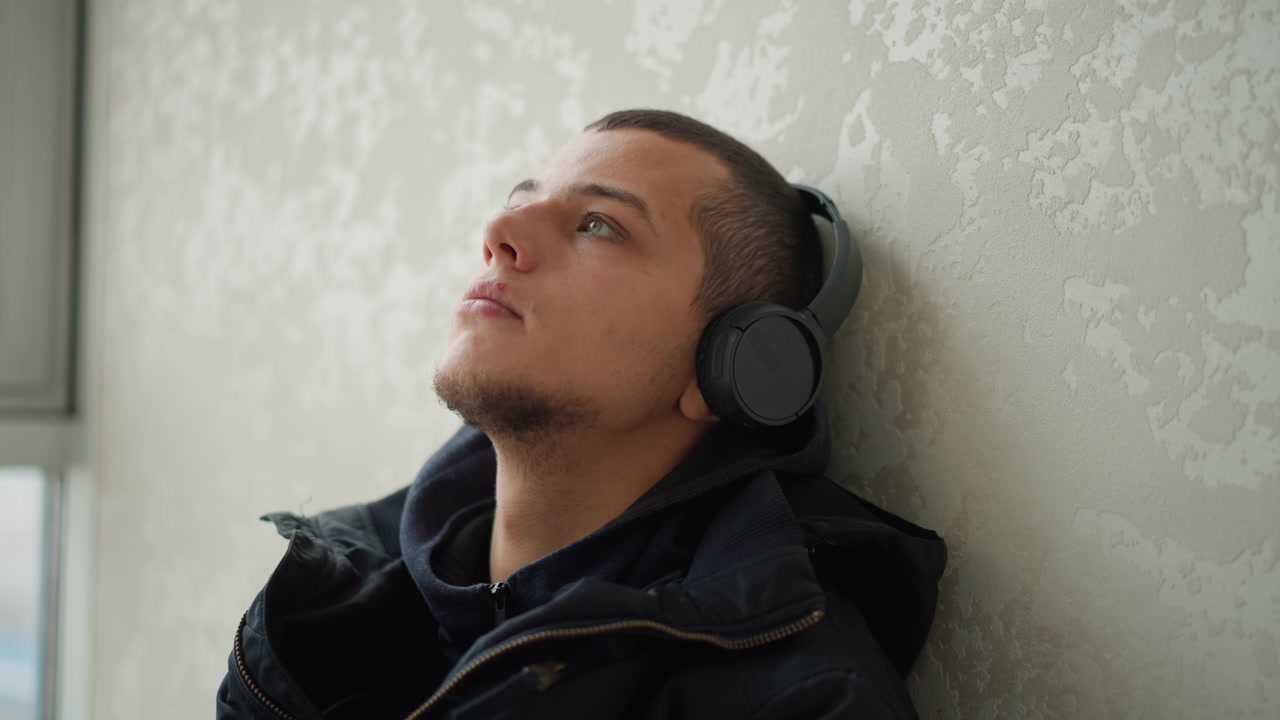 Primo piano di un uomo che indossa cuffie, appoggiato a una parete strutturata con una finestra sullo sfondo. Il soggetto rimane immobile per tutto il tempo, mantenendo un'espressione calma mentre ascolta musica.