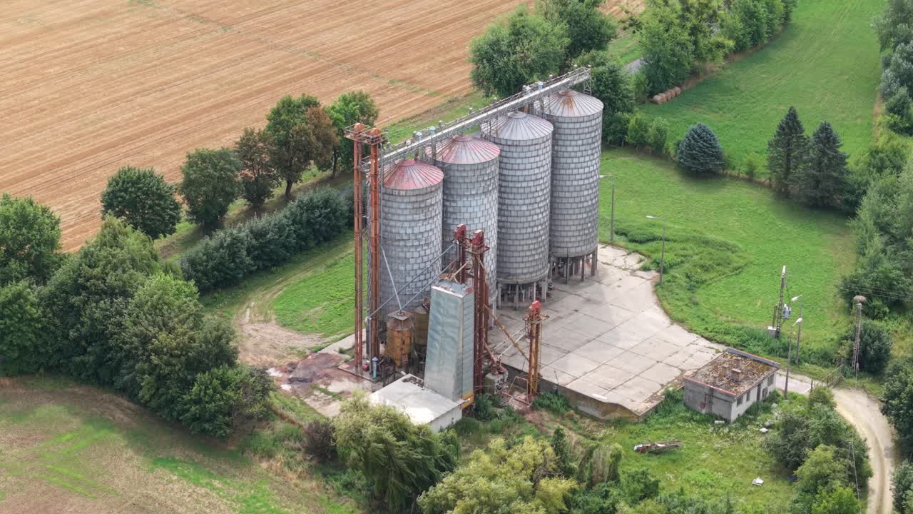 A group of large metal grain silos stands on a concrete platform amidst a rural farming landscape. The area is surrounded by green trees and cultivated fields