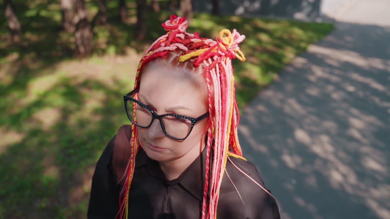 mujer caucásica melancólica con trenzas rosas y gafas caminando por una avenida arbolada y sombreada, ángulo superior, luz solar moteada y sombras suaves, retrato de un viaje solitario que transmite una soledad silenciosa
