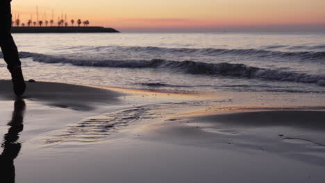 Silueta-De-Las-Piernas-De-Los-Hombres-Caminando-En-La-Playa-Durante-El-Amanecer,-Saltando-Sobre-Un-Charco-Cerca-De-La-Costa