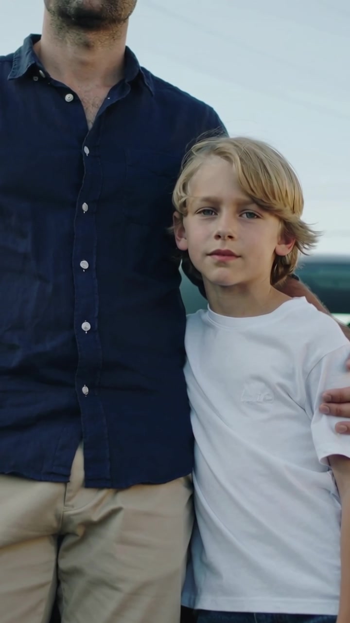 Vertikal video: Far holder sønnens skulder og poserer ved en bil, iført navyblå skjorte og hvid T-shirt.