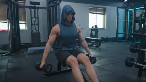 Hombre-Atlético-Haciendo-Levantamientos-Laterales-Con-Mancuernas-Sentado-En-Un-Banco-En-Un-Gimnasio-Vacío,-Usando-Pantalones-Cortos-Y-Camiseta-Sin-Mangas