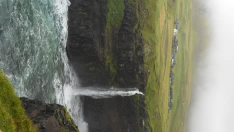 vertical - mulafossur waterfall and misty village of gasadalur on vagar island, faroe islands. static shot