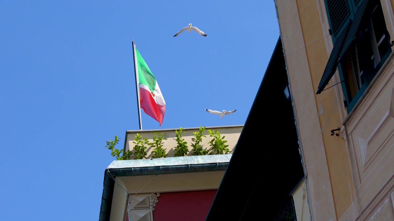 Inquadratura dal basso di una bandiera nazionale che sventola sopra un edificio mediterraneo con gabbiani