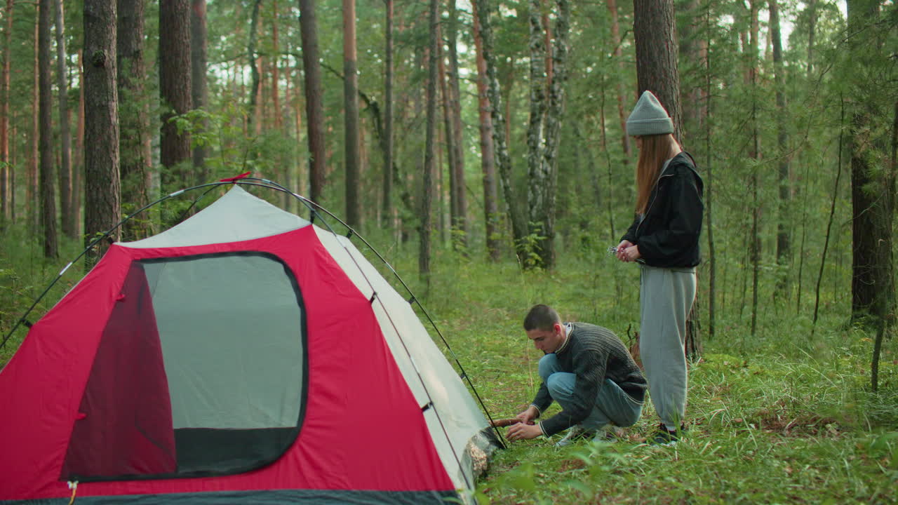 man squats beside tent hammering peg into ground with stick while woman stands beside handing him additional pegs as they set up tent together surrounded by green forest trees