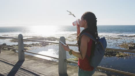 mujer afroamericana bebiendo café y usando un teléfono inteligente en el paseo marítimo