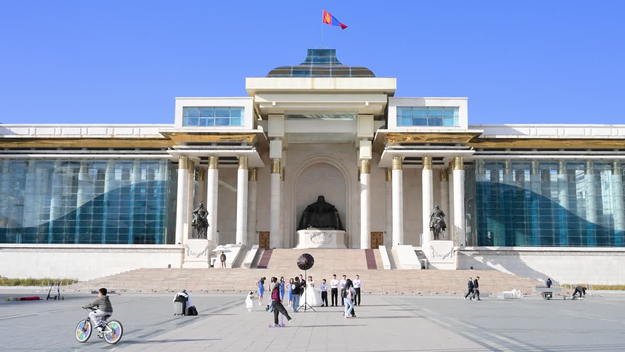 A broad view of the Government Palace at Sukhbaatar Square, Ulaanbaatar, featuring the iconic Mongolian flag and people visiting the Genghis Khan statue.