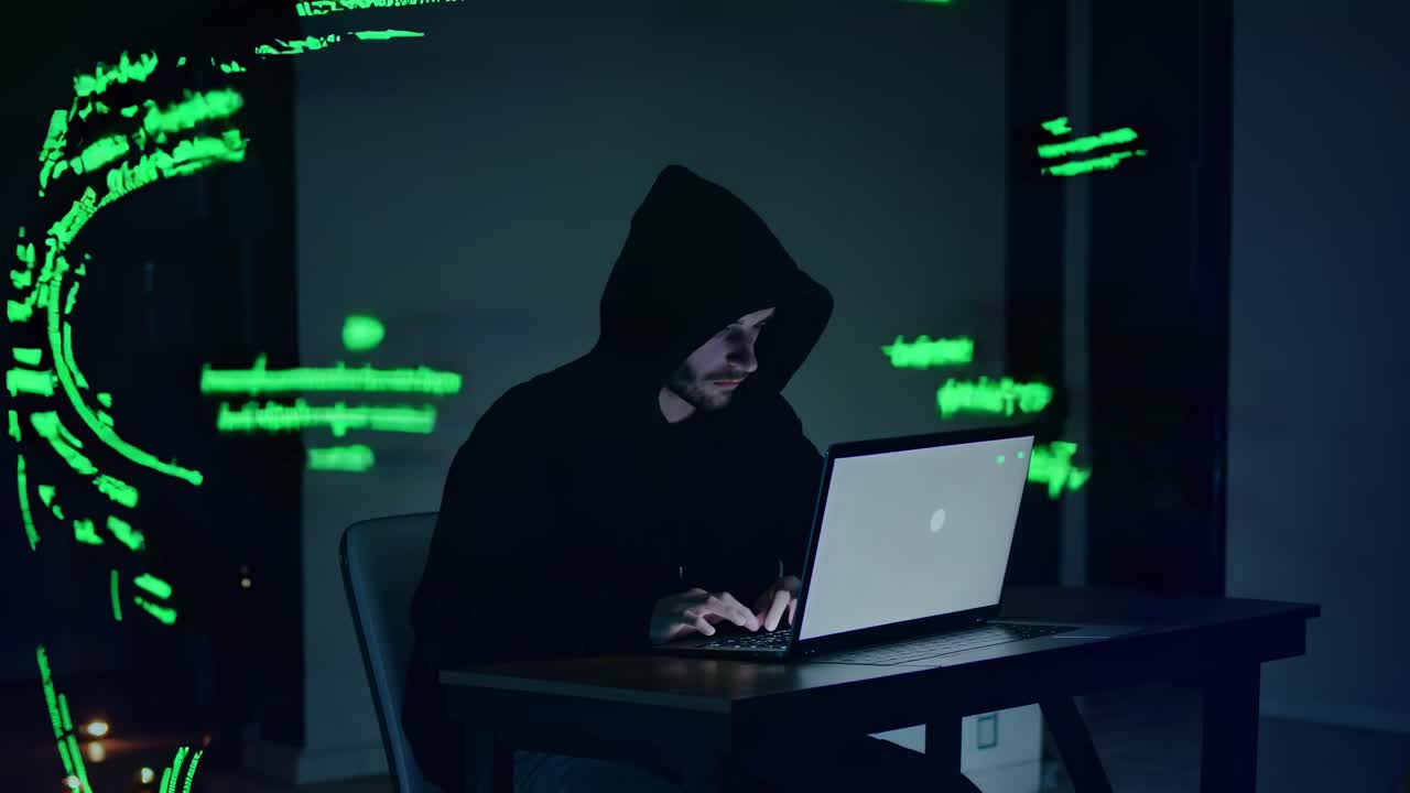 A hacker in a dark room, wearing a hoodie, types on a laptop