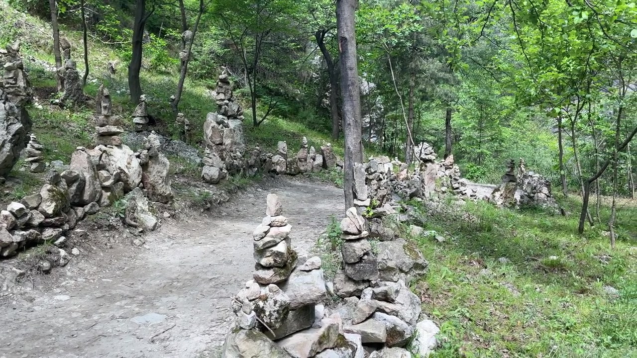 Numerous rock cairns line a dirt path through a lush green forest on a sunny day in Gunibsky District, Republic of Dagestan, Russia
