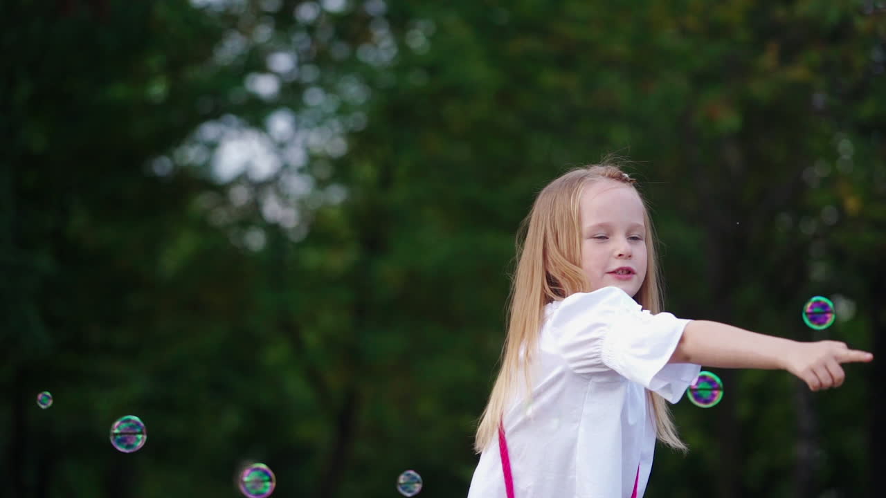 Positive little girl with soap bubbles outdoors. Smiling lovely child with blond hair popping colorful air bubbles in the park. Happy childhood.