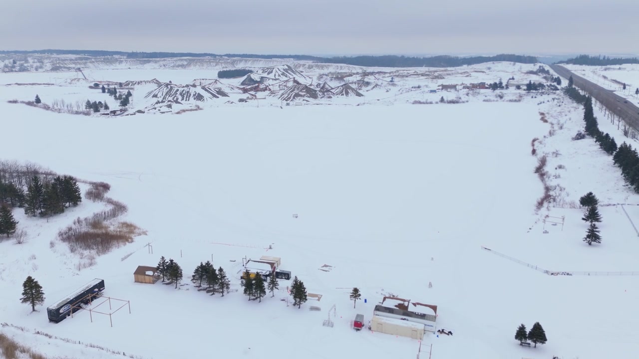 Wide aerial drone footage sweeping over a frozen gravel pit and rural Caledon landscape, Ontario, in deep winter, with snow fields, rustic buildings, winding roads, moody overcast sky.
