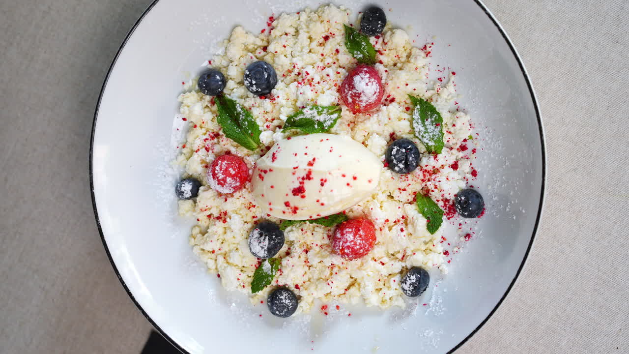 Sweet cottage cheese dessert, decorated with fresh blueberries, raspberries, mint leaves, and powdered sugar, is presented on a white plate