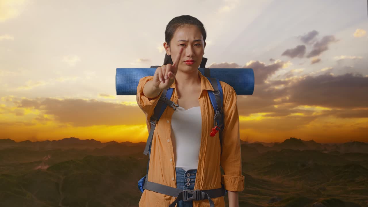 excursionista asiática con mochila de montañismo que no muestra ningún gesto con el dedo índice mientras está de pie en la cima de la montaña durante la hora del atardecer