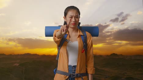 asian female hiker with mountaineering backpack showing no gesture with index finger while standing on the top of mountain during sunset time