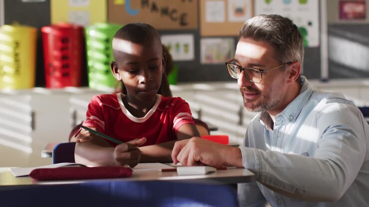 Diverse happy male teacher helping schoolboy sitting in classroom during learning