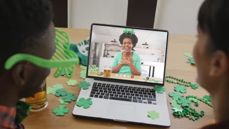 Smiling-african-american-woman-with-beer-wearing-clover-shape-band-on-video-call-on-laptop