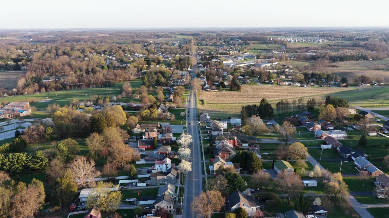 Vista aerea con drone di un'area suburbana della Pennsylvania con una strada principale dritta, circondata da case, alberi e terreno aperto sotto la calda luce solare primaverile. Paesaggio residenziale e semi-rurale calmo. Inquadratura ampia.