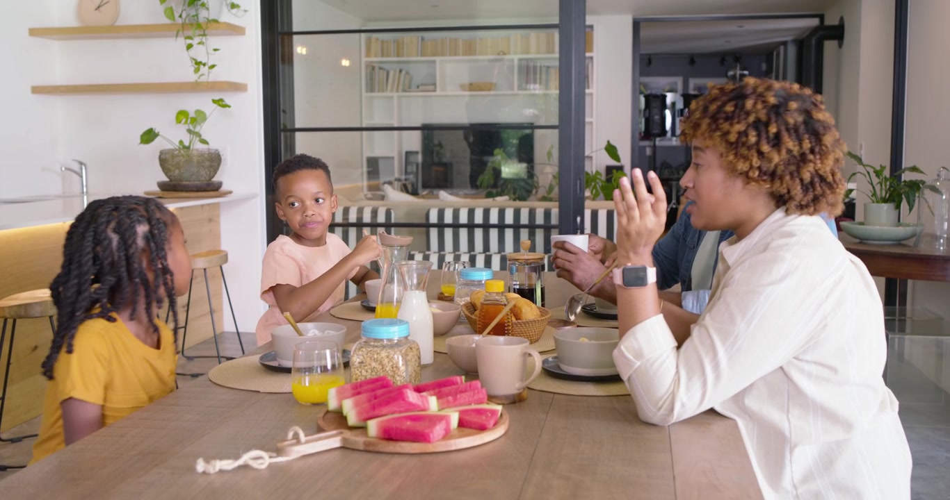 Famiglia afroamericana che fa colazione al tavolo, padre che si protende, mentre i genitori danno i cereali ai bambini.