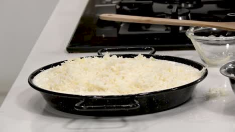 a skilled cook sprinkles grated cheese over a layer of rice in a black baking dish. the kitchen is bright and modern, showcasing culinary tools and fresh ingredients