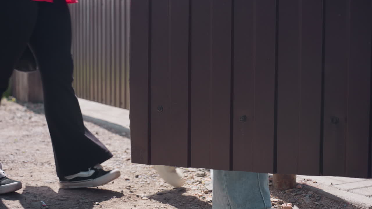 Low angle rear view shows young women in blue jeans walking dusty rural lane, one swinging paper grocery bag, other holding black tote, stride casting shadows on uneven ground during summer walk