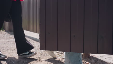 low angle rear view shows young women in blue jeans walking dusty rural lane, one swinging paper grocery bag, other holding black tote, stride casting shadows on uneven ground during summer walk