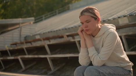 close-up of thoughtful young woman in casual hoodie and jeans sitting on rustic stadium bleachers, resting hands on chin with a pensive and reflective expression