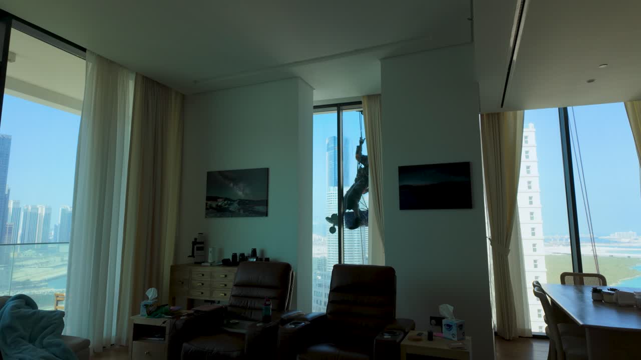 Watching from inside a skyscraper apartment as a window washer cleans the outside while rappelling from a rope