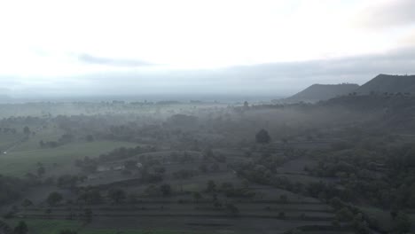 Small-forest-surrounded-by-fog-and-some-hills-on-the-background-and-it-has-some-fields-too