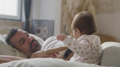 Relaxed-Married-Couple-Lying-In-Bed-On-Sunday-Morning,-Spending-Time-With-Their-Cute-Baby-Girl-Who-Playing-With-Teddy-Bear