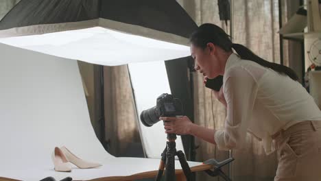 asian female photographer talking on smartphone while taking photos of women's shoes in home studio