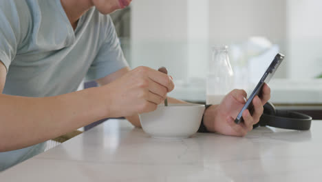 asian male teenager sitting at table with smartphone and having breakfast