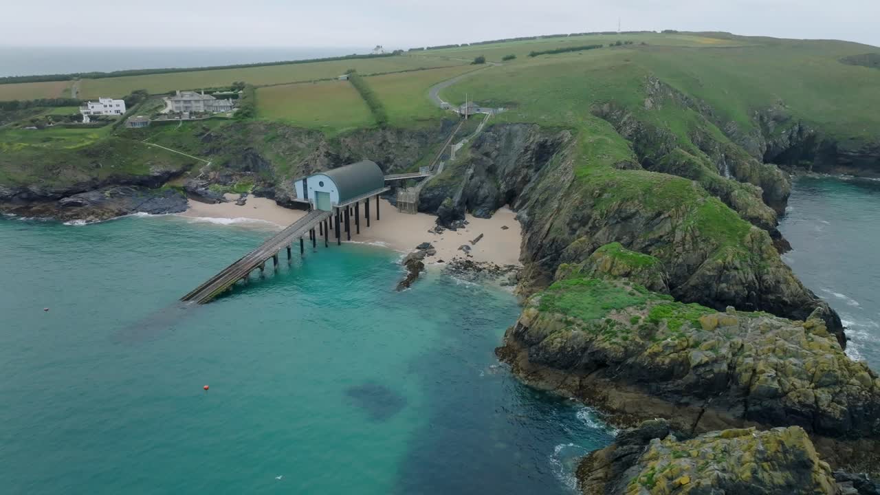 RNLI lifeboat station with long slipway next to clean sandy beach with clear blue seawater. Camera ascending orbit revealing craggy sea cliffs. Mother Ivey's Bay, Cornwall, UK.