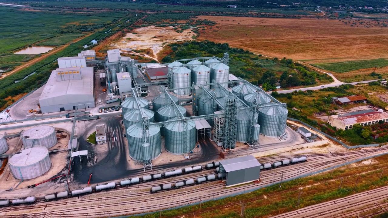 Getreideverarbeitungsanlage mit Gleisanschluss. Schräge Luftaufnahme von Metallsilos, Fabrikhallen, Fördertürmen, Kesselwagen und umliegenden Feldern.