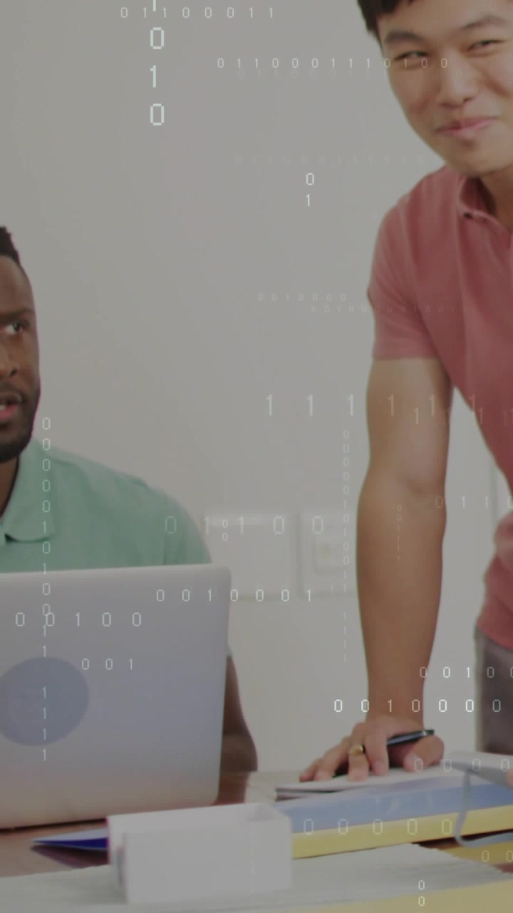 Vertical video: Seated man typing at laptop while standing man leaning in showing tech data overlay