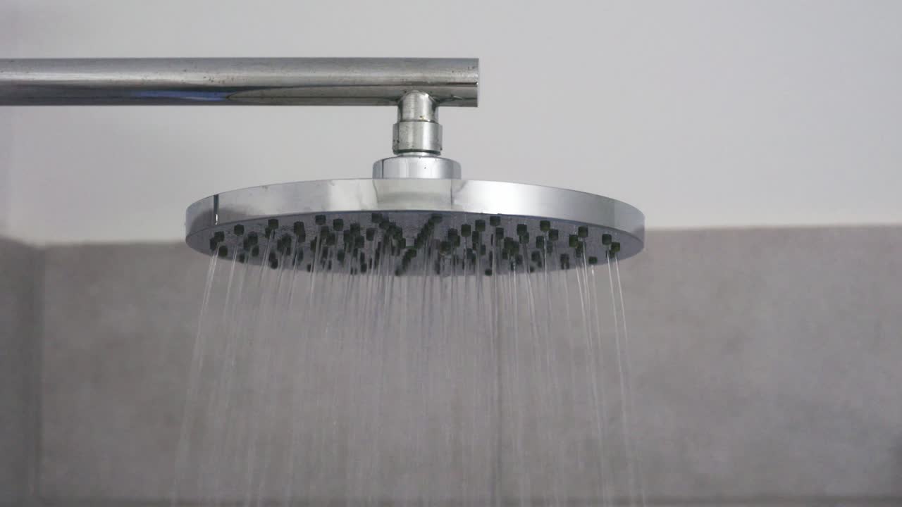 A modern chrome shower head releases a steady stream of water in a clean, minimal bathroom setting. The close-up angle highlights the sleek metallic design and the gentle downward flow of water