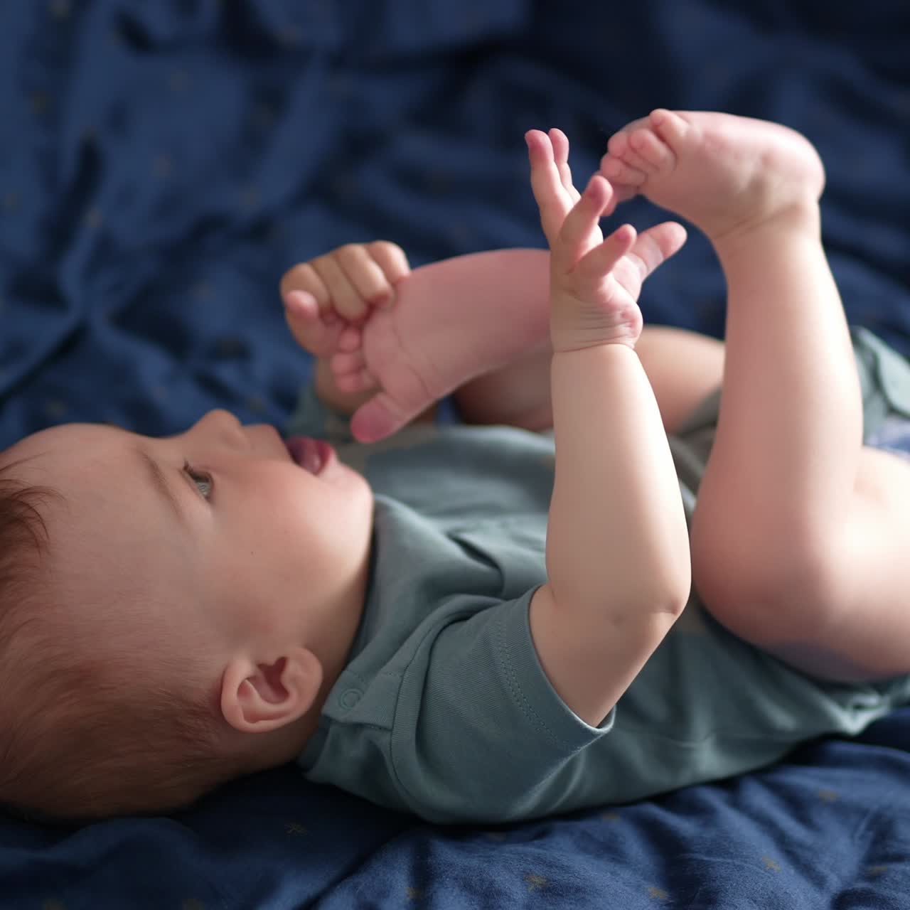 Adorable Caucasian toddler lies on his back on the bed. Beautiful child plays with his feet pulling them to mouth