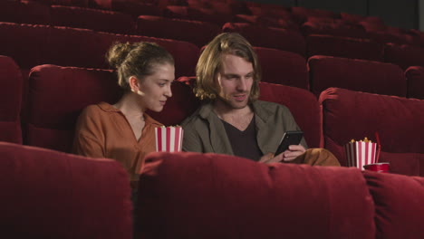 hombre rubio sentado en el cine y usando un teléfono móvil mientras su novia come palomitas de maíz
